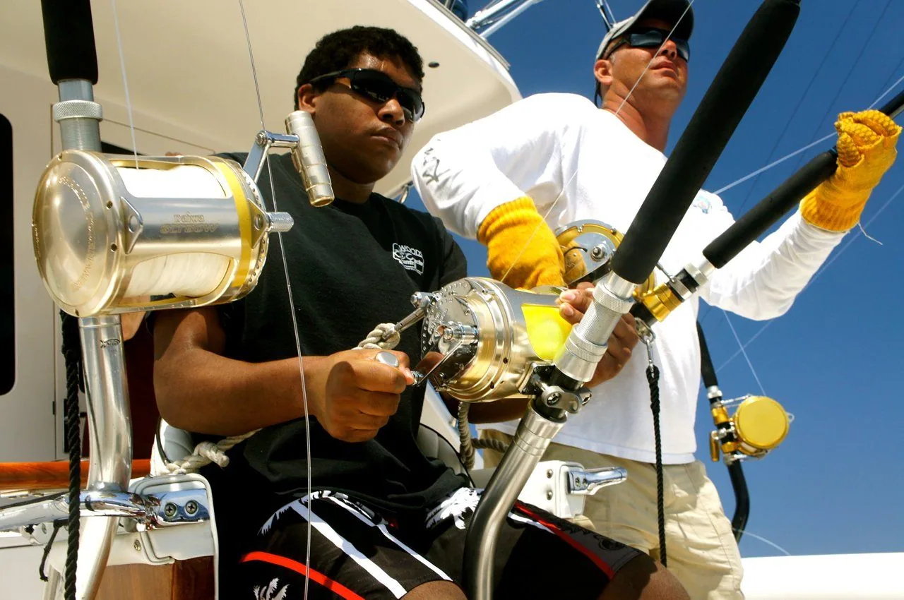 Two men are fishing on a boat and one of them is wearing a black shirt that says adidas
